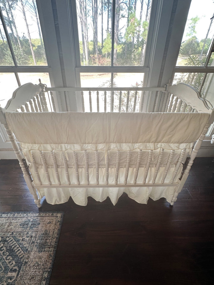 Backlit front view of the ivory ruffled rail cover across the top rail, farmhouse skirt gathered to dark wood floors