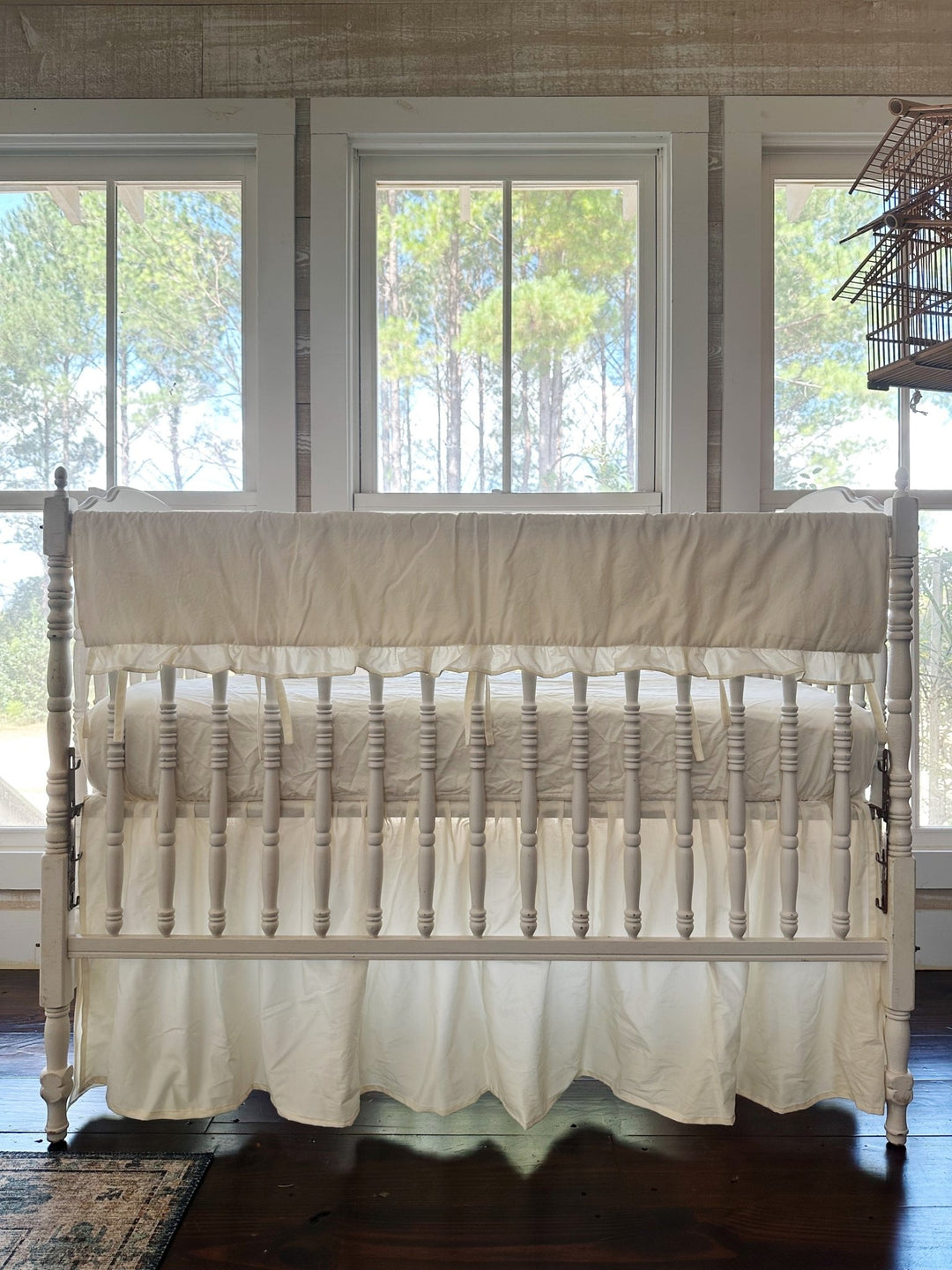 Ivory ruffled crib rail cover and farmhouse crib skirt on a white spindle crib, straight-on in front of three tall windows
