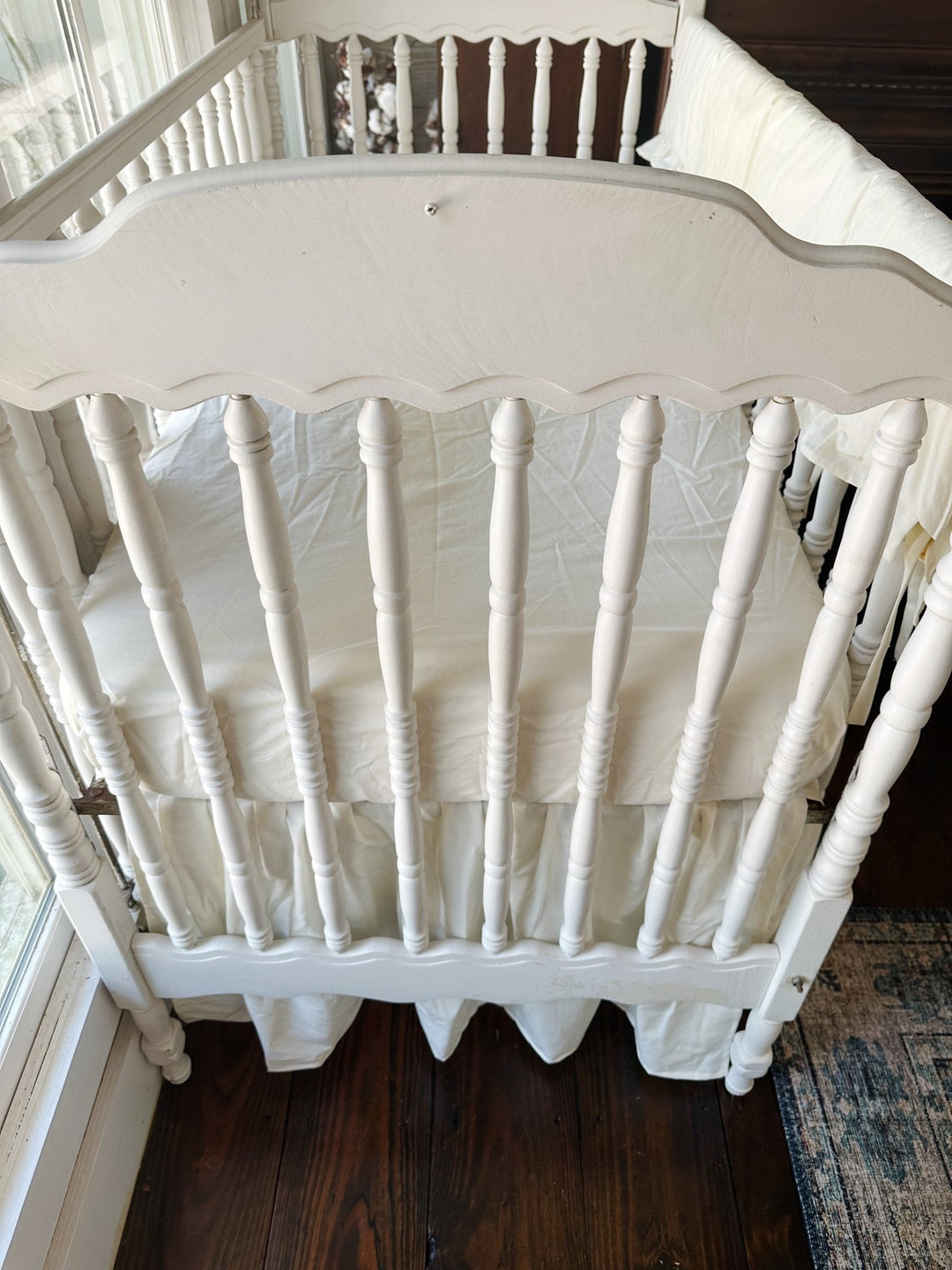 Looking down over the scalloped end of the crib at an ivory fitted mattress, gathered ivory skirt below and the rail cover along the side
