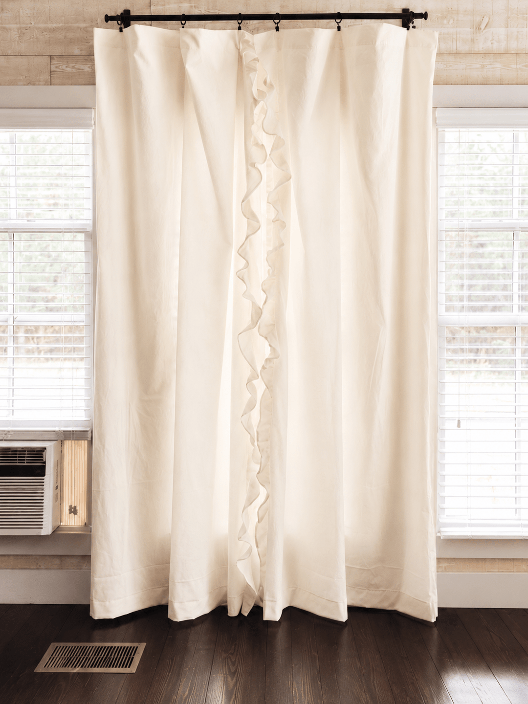 Ivory ruffled nursery curtains hung on a black rod across a window, panels falling to dark wood floors - High Cotton Textile