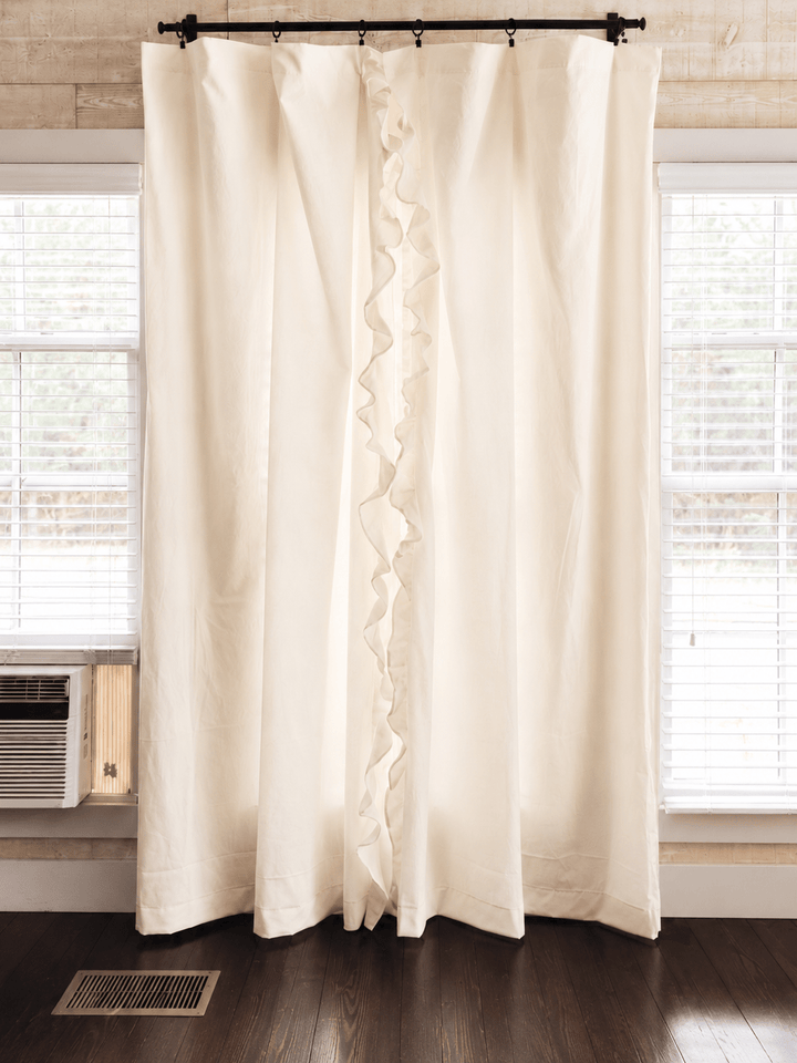 Ivory ruffled nursery curtains hung on a black rod across a window, panels falling to dark wood floors - High Cotton Textile