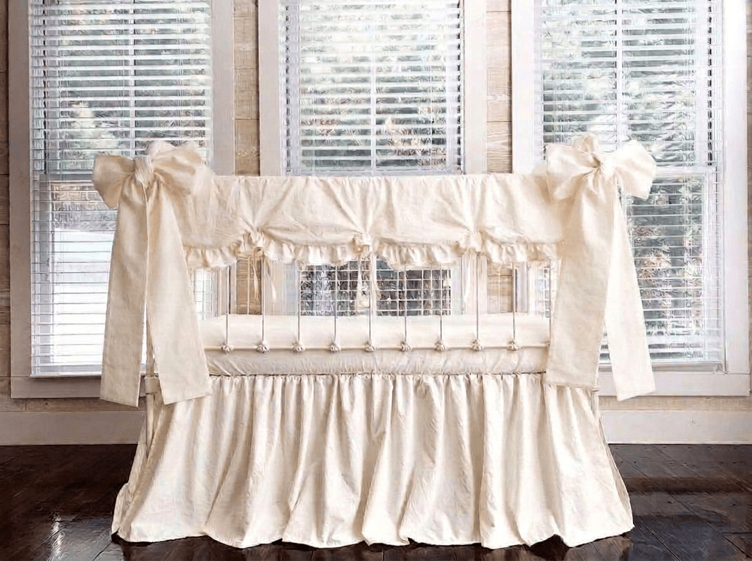 Ivory scalloped crib rail cover, ivory farmhouse skirt and two large ivory crib bows tied at the front corner posts of a white iron crib