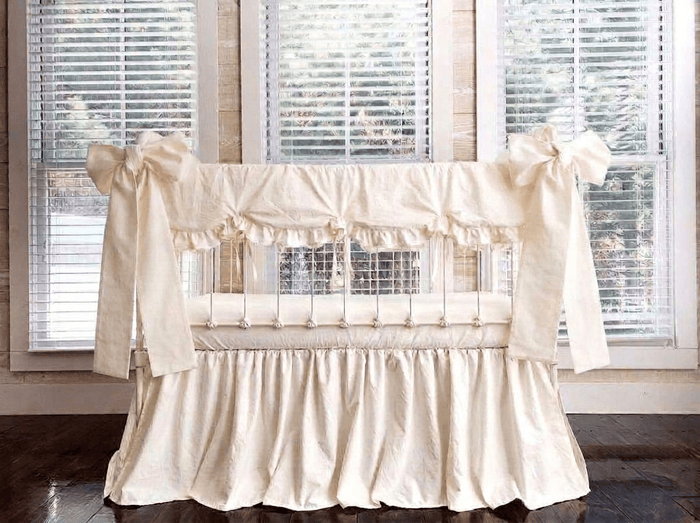 Ivory scalloped crib rail cover, ivory farmhouse skirt and two large ivory crib bows tied at the front corner posts of a white iron crib