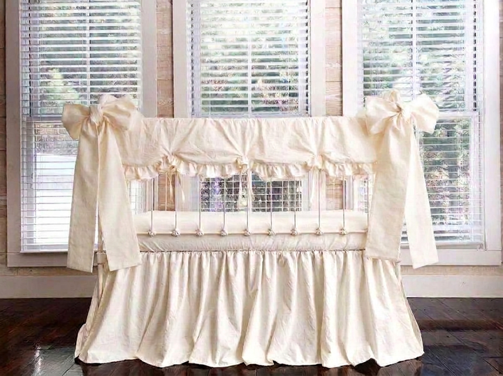 Ivory scalloped crib rail cover, ivory farmhouse skirt and two large ivory crib bows tied at the front corner posts of a white iron crib