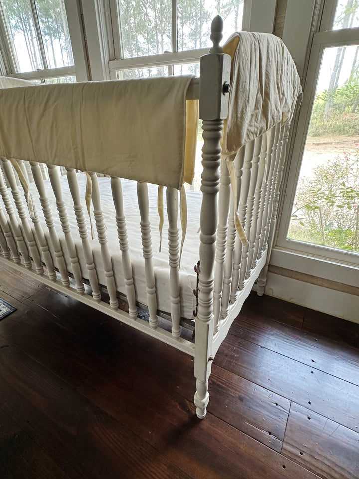 Ivory crib rail covers seen from the corner of the crib with windows and greenery behind