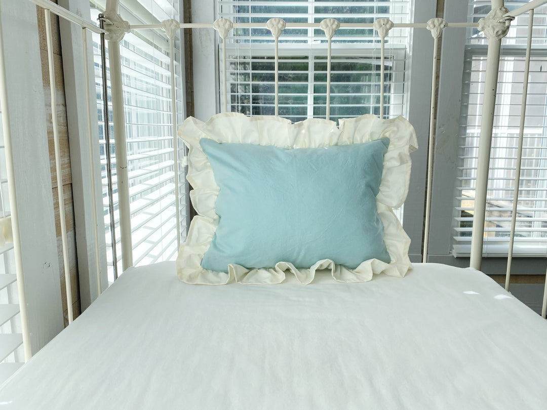 Ivory ruffled crib pillow in mist blue shown inside a white iron crib