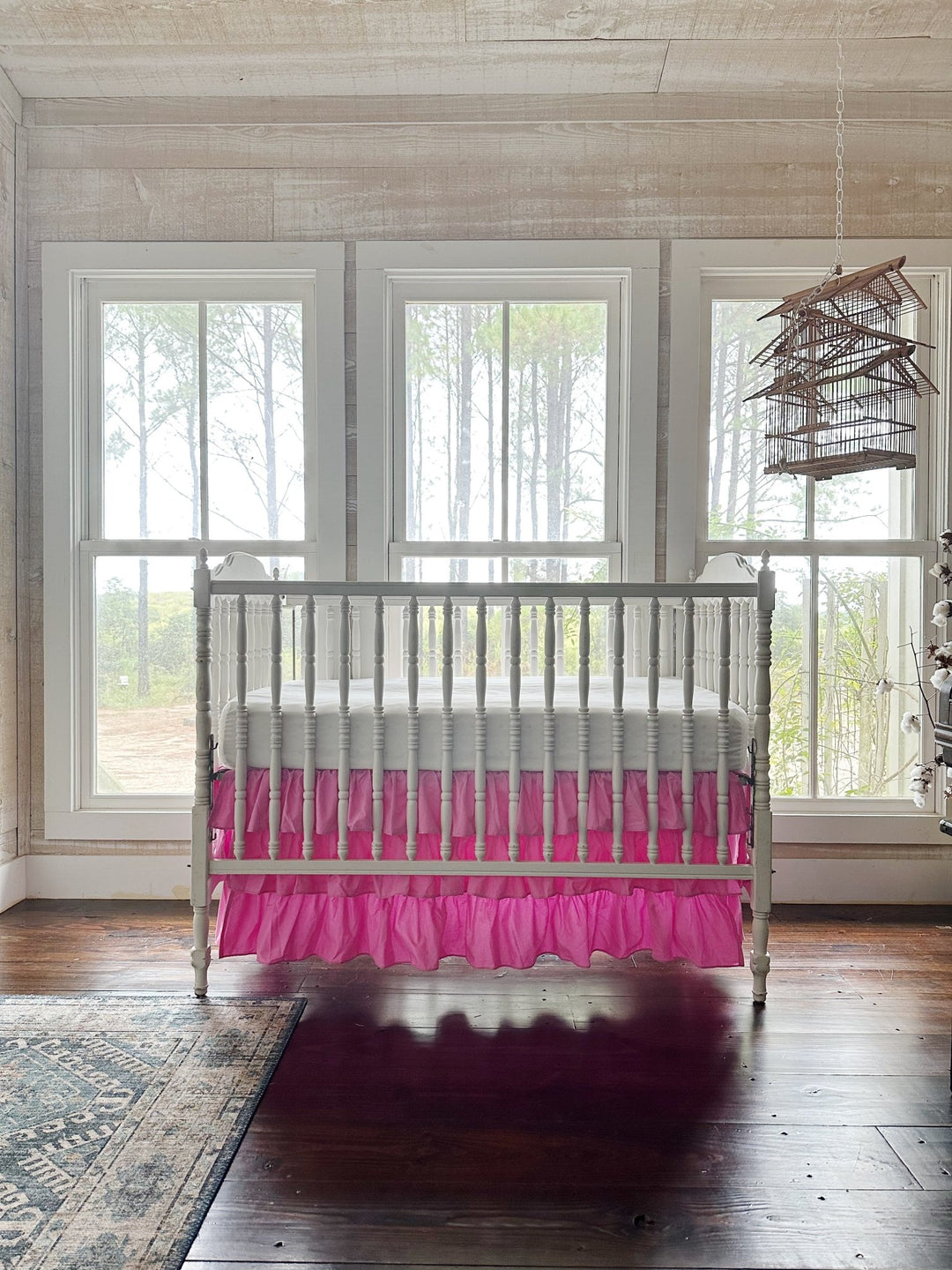 Nursery with whitewashed plank walls and a hanging birdcage, white spindle crib dressed in a pink three-tier ruffled crib skirt