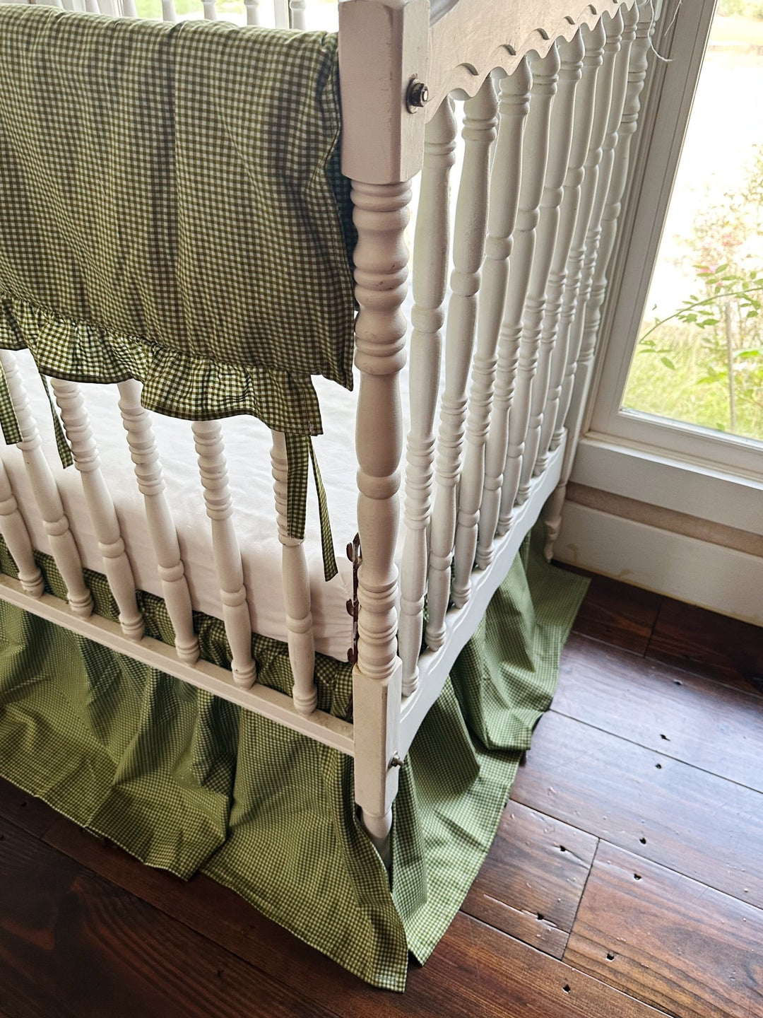 Corner detail of green gingham crib rail cover tied to a white crib rail above the crib skirt