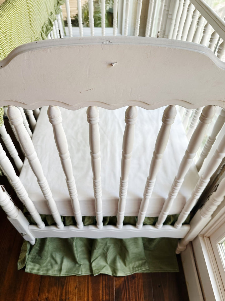 End view of white spindle crib with green gingham crib skirt and white crib sheet