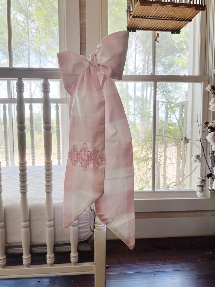 Pink buffalo check crib bow tied to a white spindle crib post with a pink monogram on the tail
