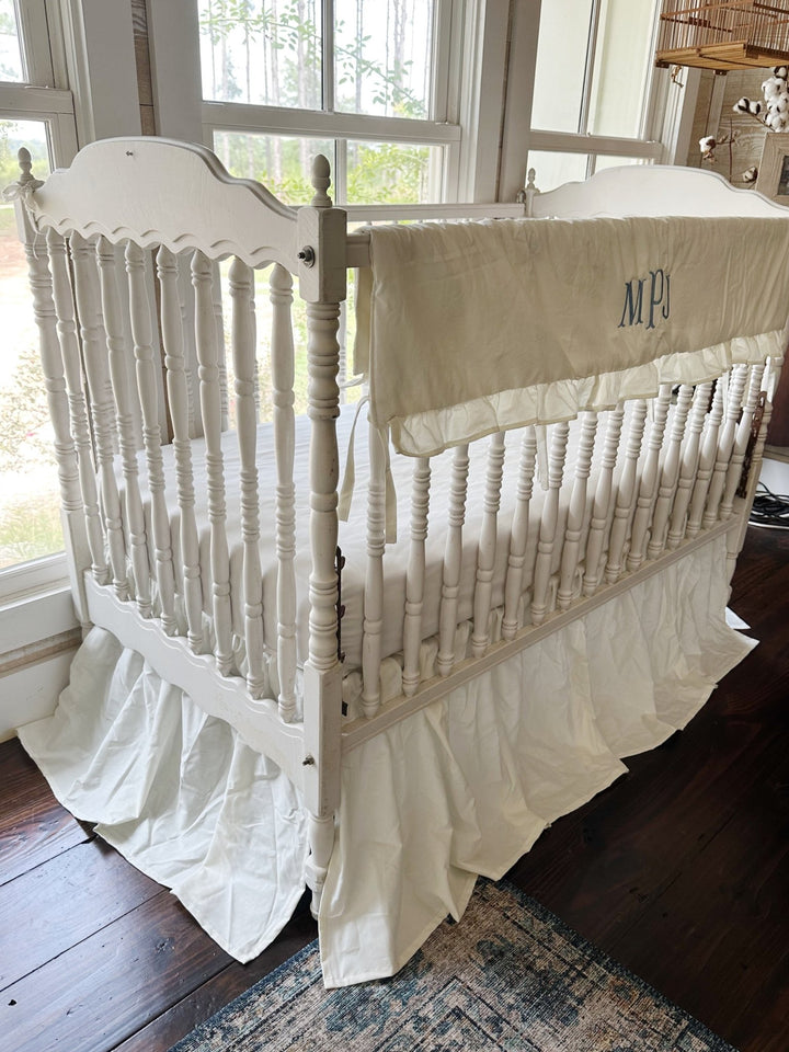 Three-quarter view of the monogrammed rail cover with the skirt gathered around the crib legs
