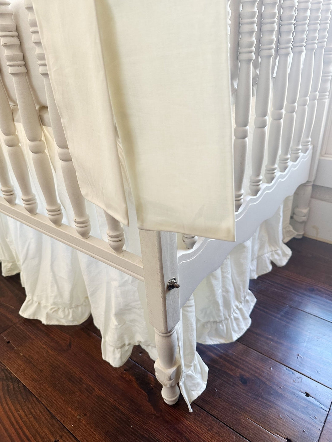 Low corner view of a porcelain bow tail beside the crib leg, ruffled skirt hem pooling on the wood floor