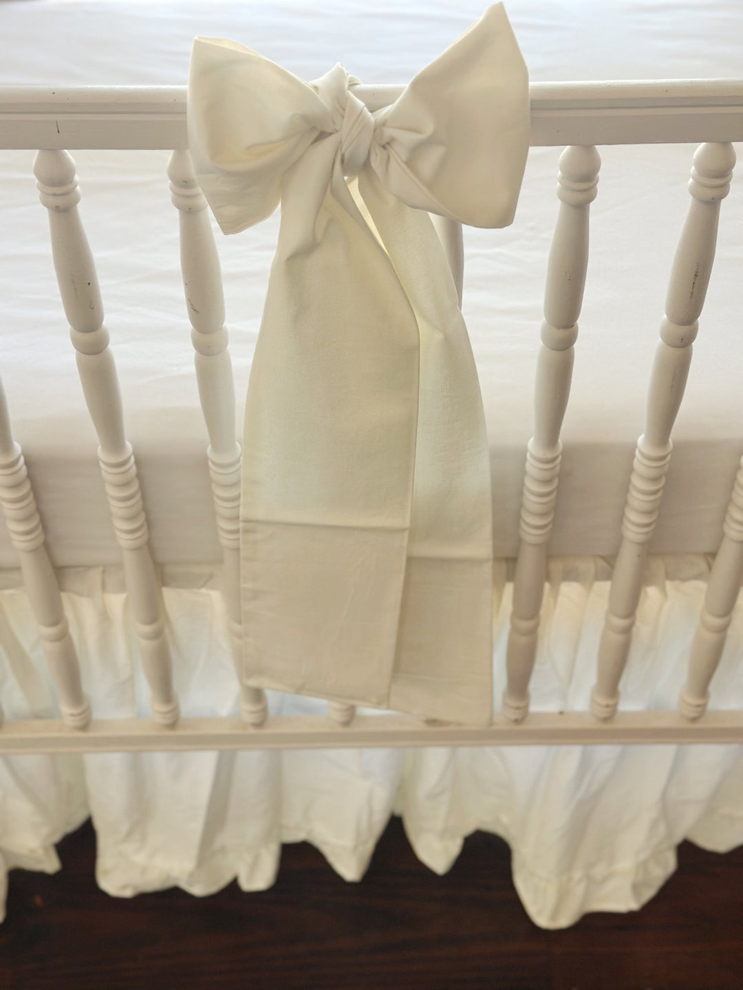 Close-up of a porcelain cotton bow knotted at the center of the front crib rail, tails falling over the gathered skirt