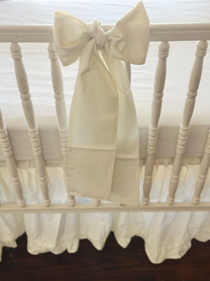 Close-up of a porcelain cotton bow knotted at the center of the front crib rail, tails falling over the gathered skirt