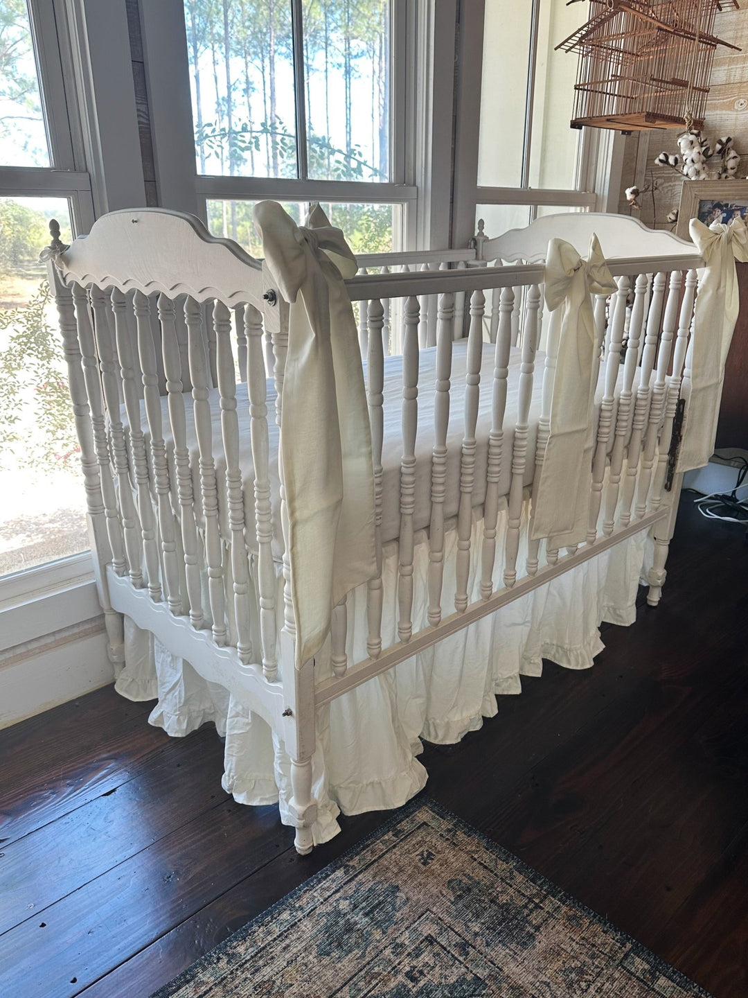 Three-quarter view of the porcelain ruffled crib skirt and bows on a white spindle crib by the nursery windows