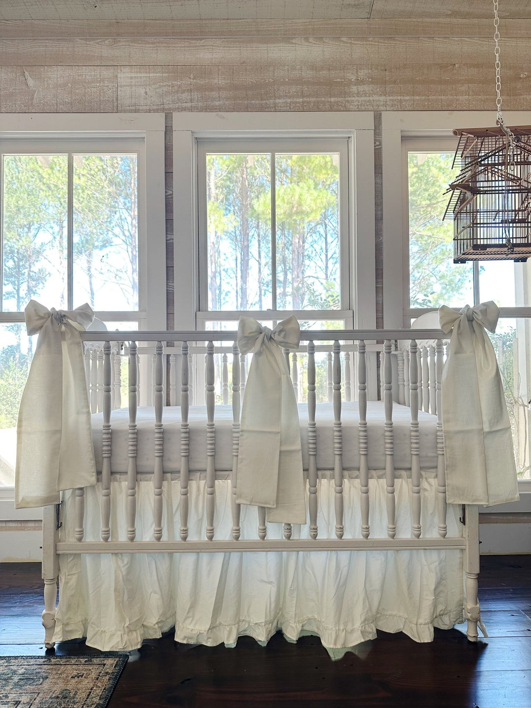 Porcelain ruffled crib skirt and three porcelain crib bows on a white spindle crib, front view in front of three tall windows