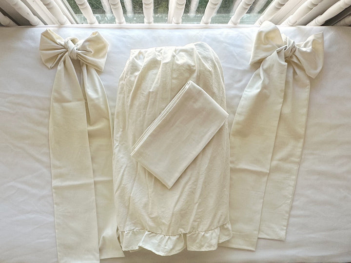 Porcelain ruffled crib skirt panel laid flat between two tied porcelain crib bows with long sash tails