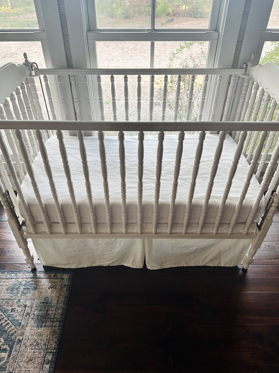 Porcelain crib skirt with its pleated center panel hanging below the crib rail in front of a sunlit window - High Cotton Textile