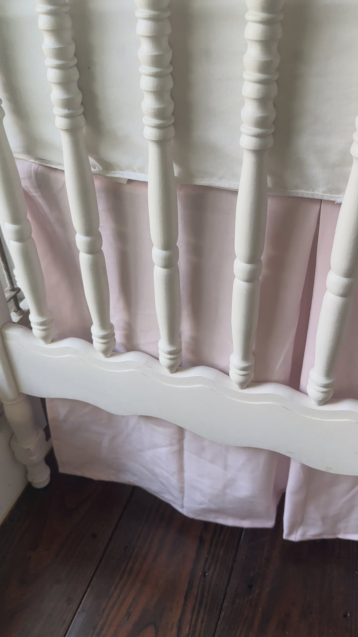 Video of the baby pink tailored cotton crib skirt on a white spindle crib, showing the pleats and split corners