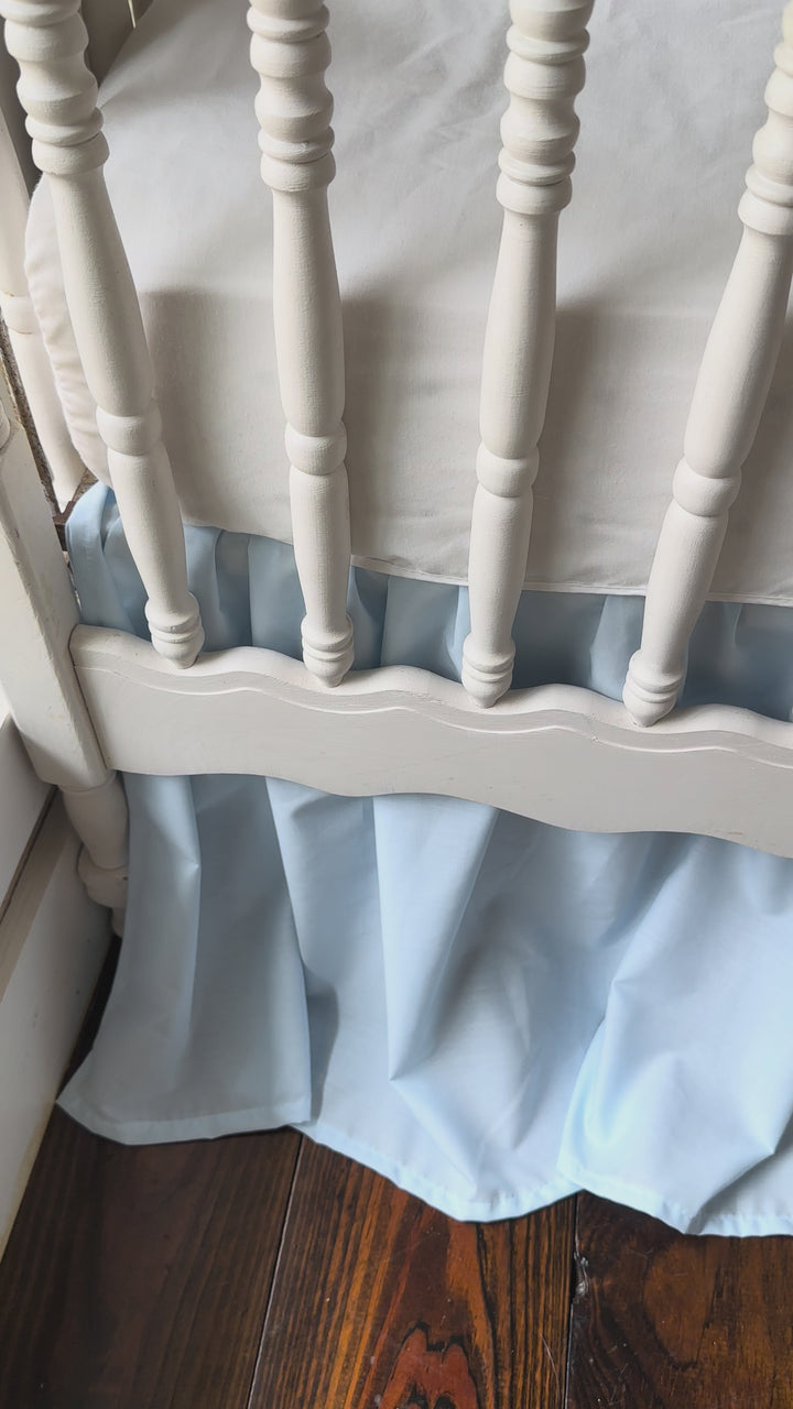 White scalloped crib rail cover with heirloom blue ruffle and matching farmhouse crib skirt on a spindle crib.