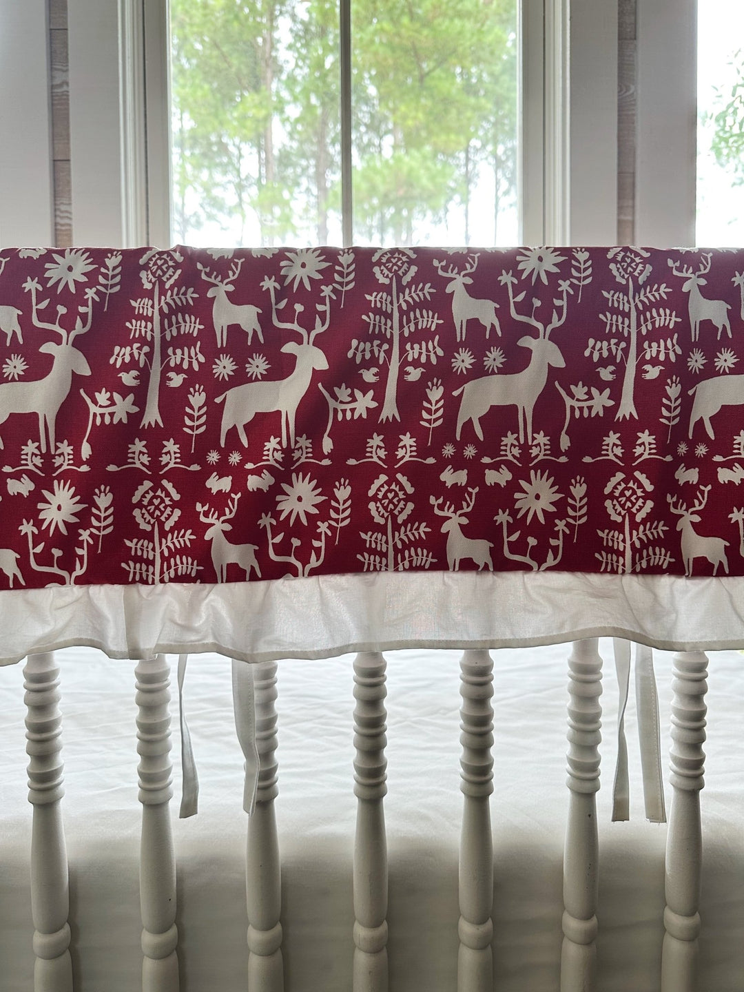 Red and white folk deer ruffled crib rail cover on the long rail of a white spindle crib