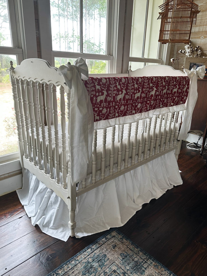 Red and white folk deer ruffled crib rail cover with a white farmhouse skirt and two large white bows on a spindle crib