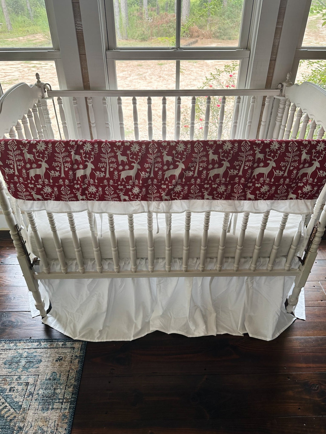 Cranberry deer print rail cover and white gathered skirt on a white spindle crib, seen from above