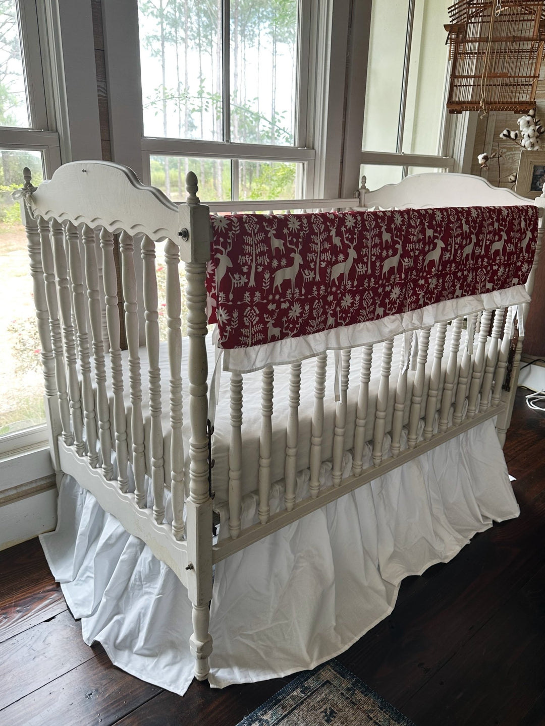 Red and white folk deer ruffled crib rail cover above a white farmhouse crib skirt, seen from the corner