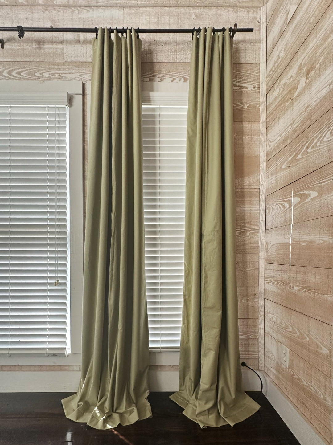 Pair of sage green blackout curtains pulled open either side of a window with white blinds, hems breaking on dark floorboards
