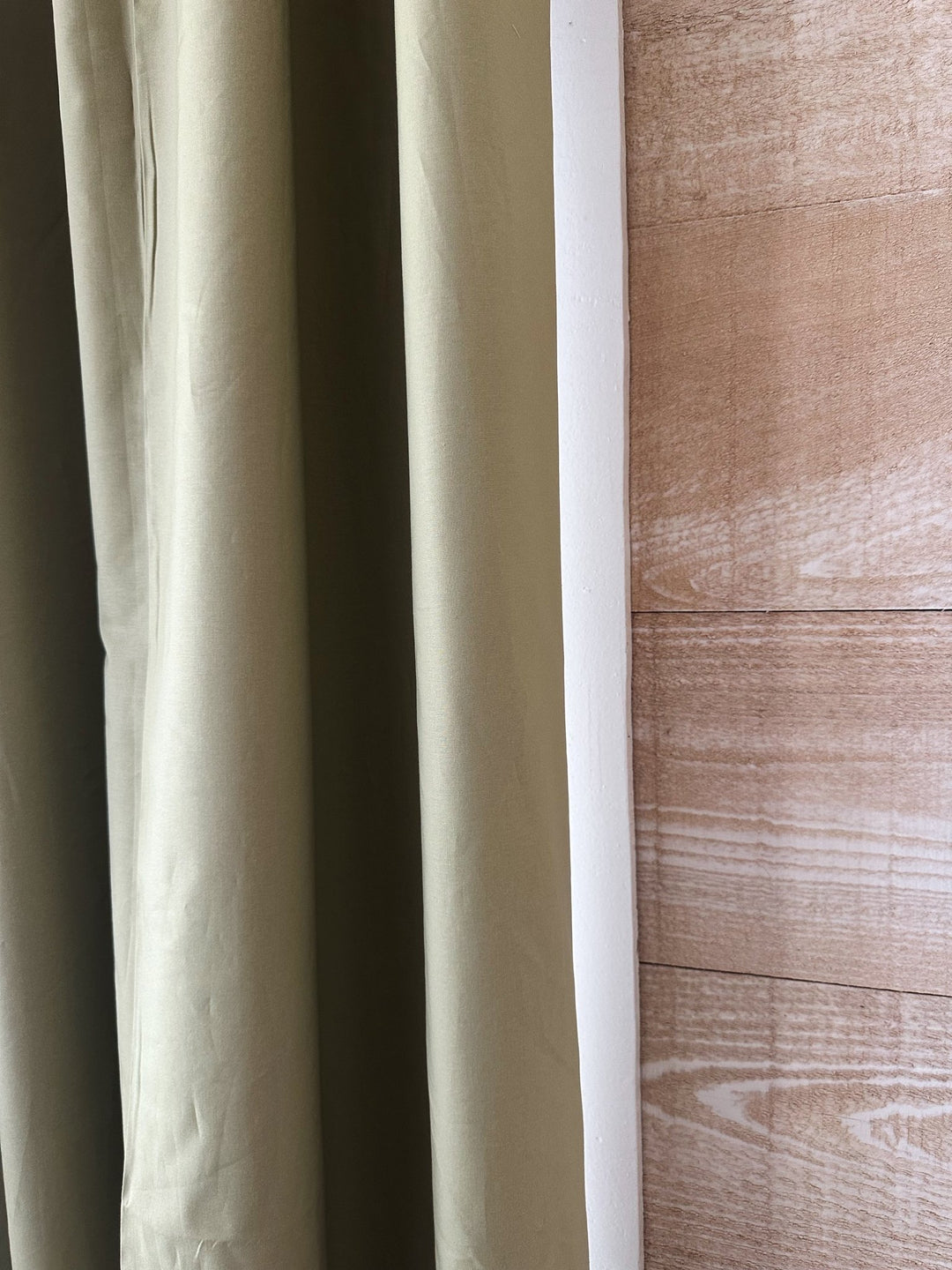 Edge of a sage green cotton panel hanging beside white window trim and a warm plank wall