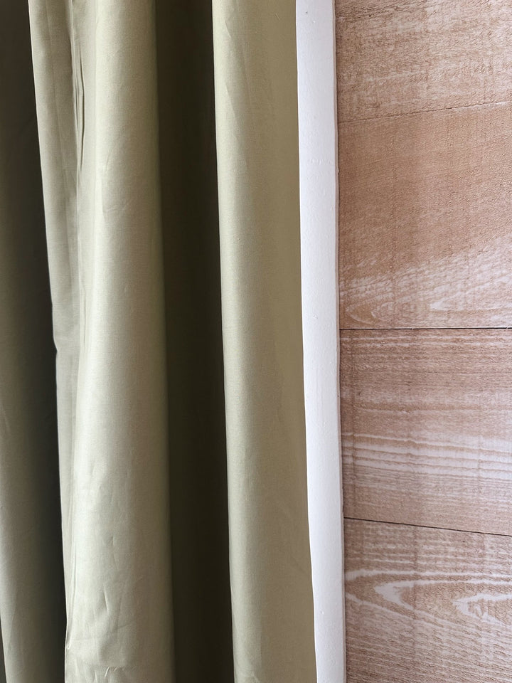 Edge of a sage green cotton panel hanging beside white window trim and a warm plank wall