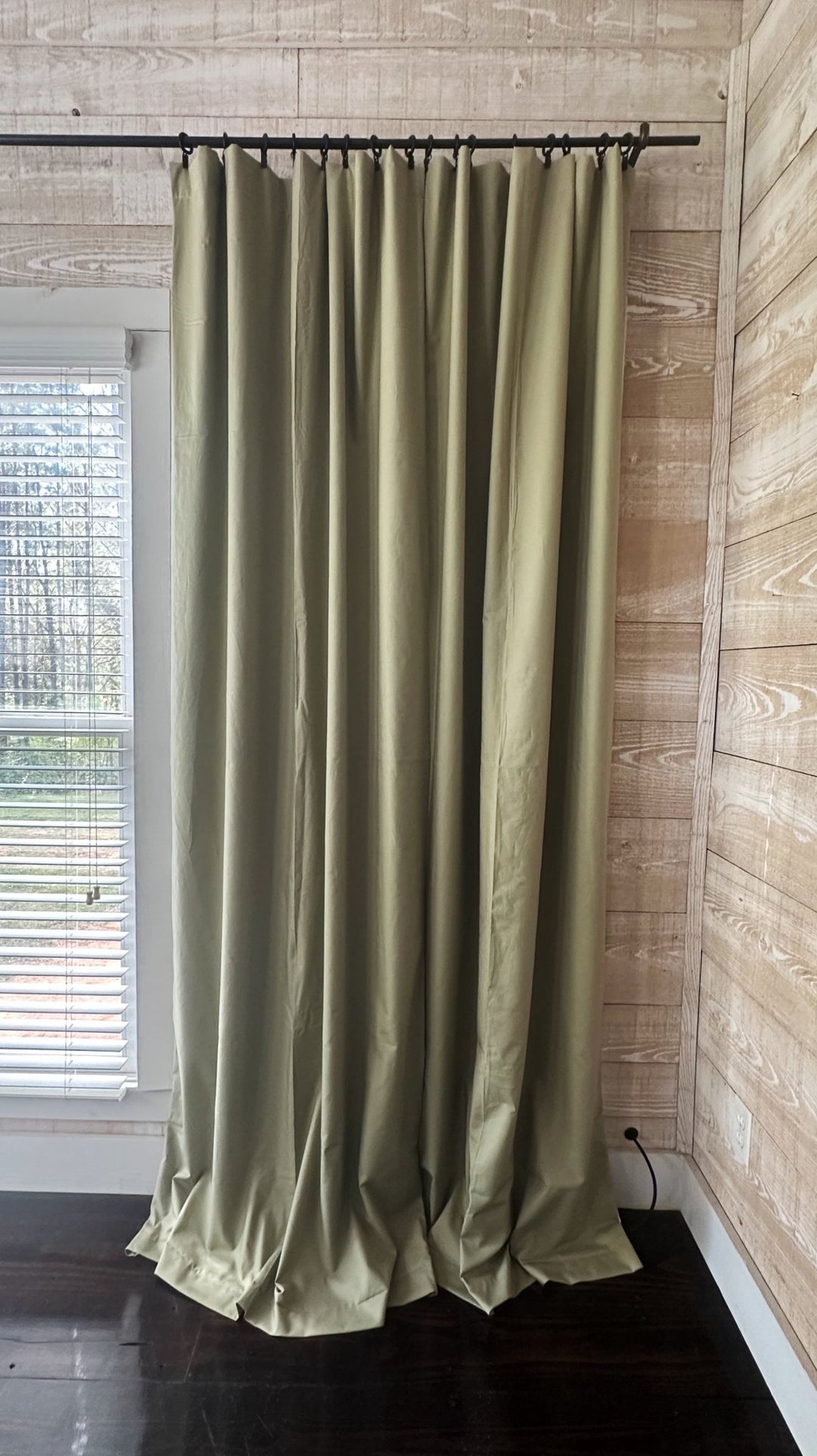 Sage green blackout panels drawn closed on a black iron rod with clip rings, rod to floor against a whitewashed plank wall