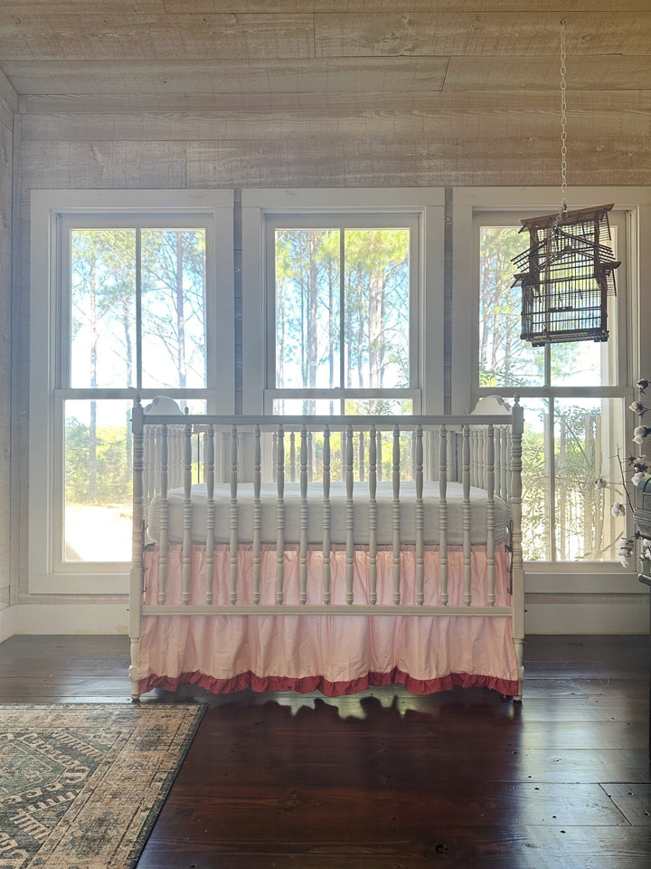 Nursery with shiplap walls and a hanging birdcage — white spindle crib dressed in a Sisters Pink crib skirt with a deeper blush ruffle hem