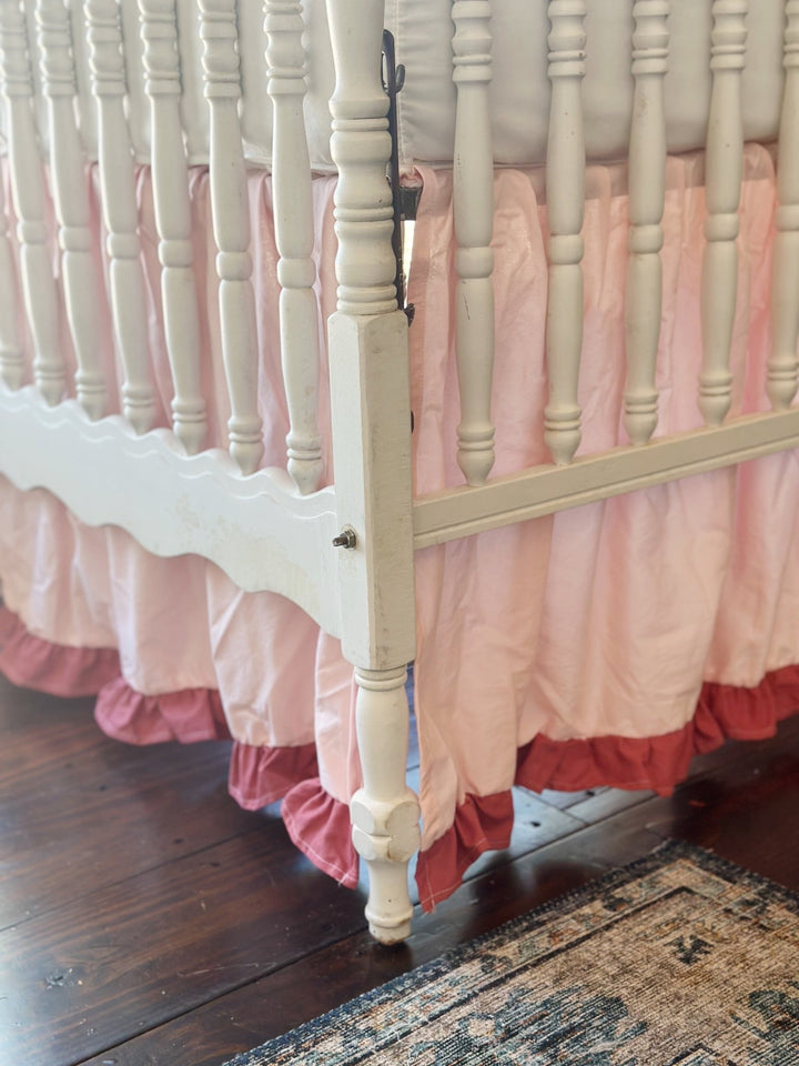 Crib corner with scalloped end rail and vintage rug, showing the pink skirt and blush ruffle turning the corner