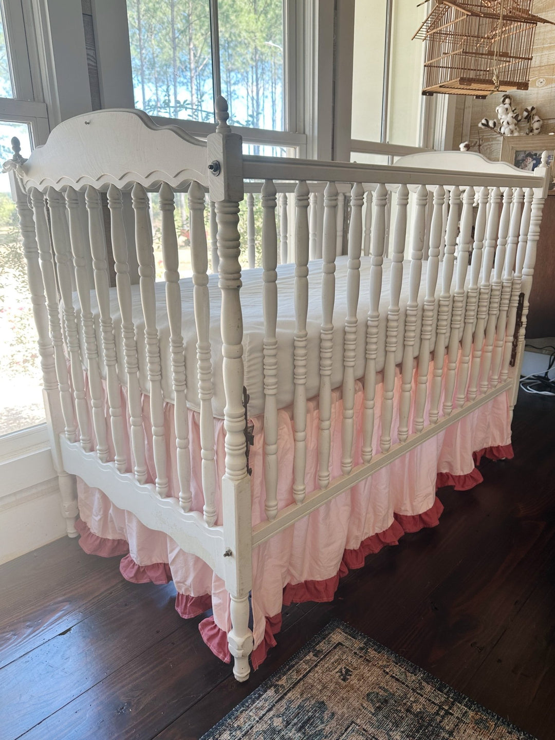 Three-quarter view of the pink ruffled crib skirt wrapping the foot end of a white spindle crib beside the windows