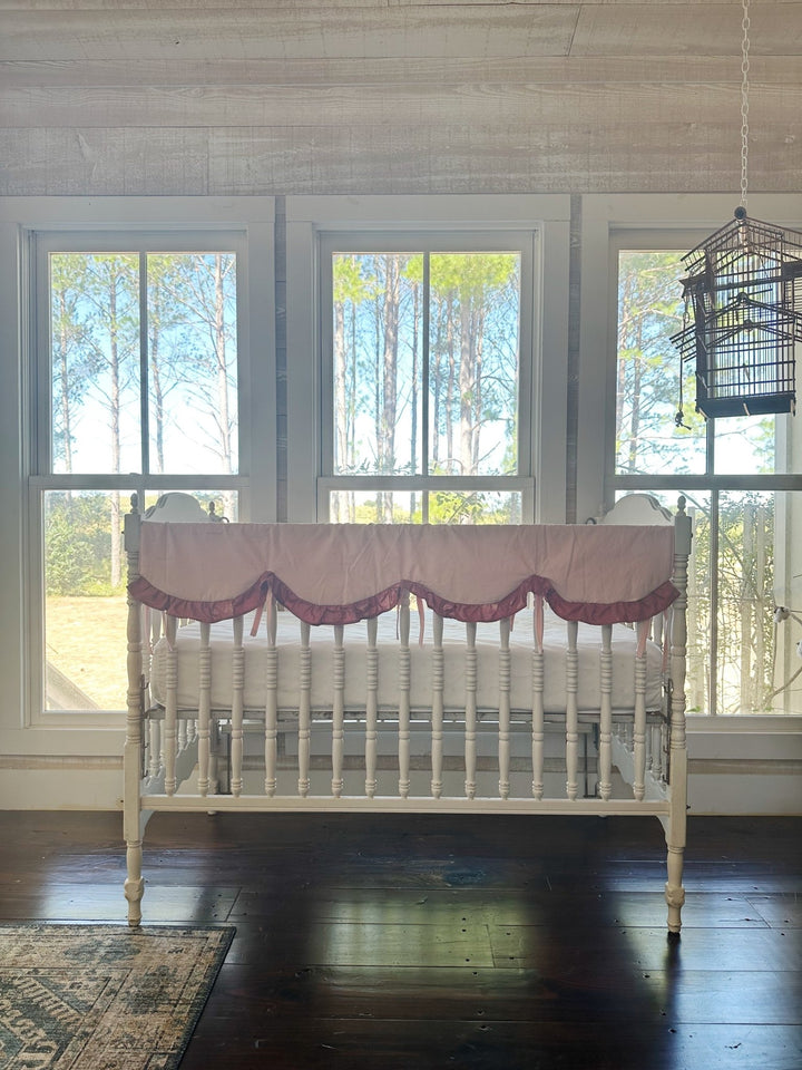 Wide nursery view of the Sisters Pink and Blush rail cover on a white crib in front of three tall windows