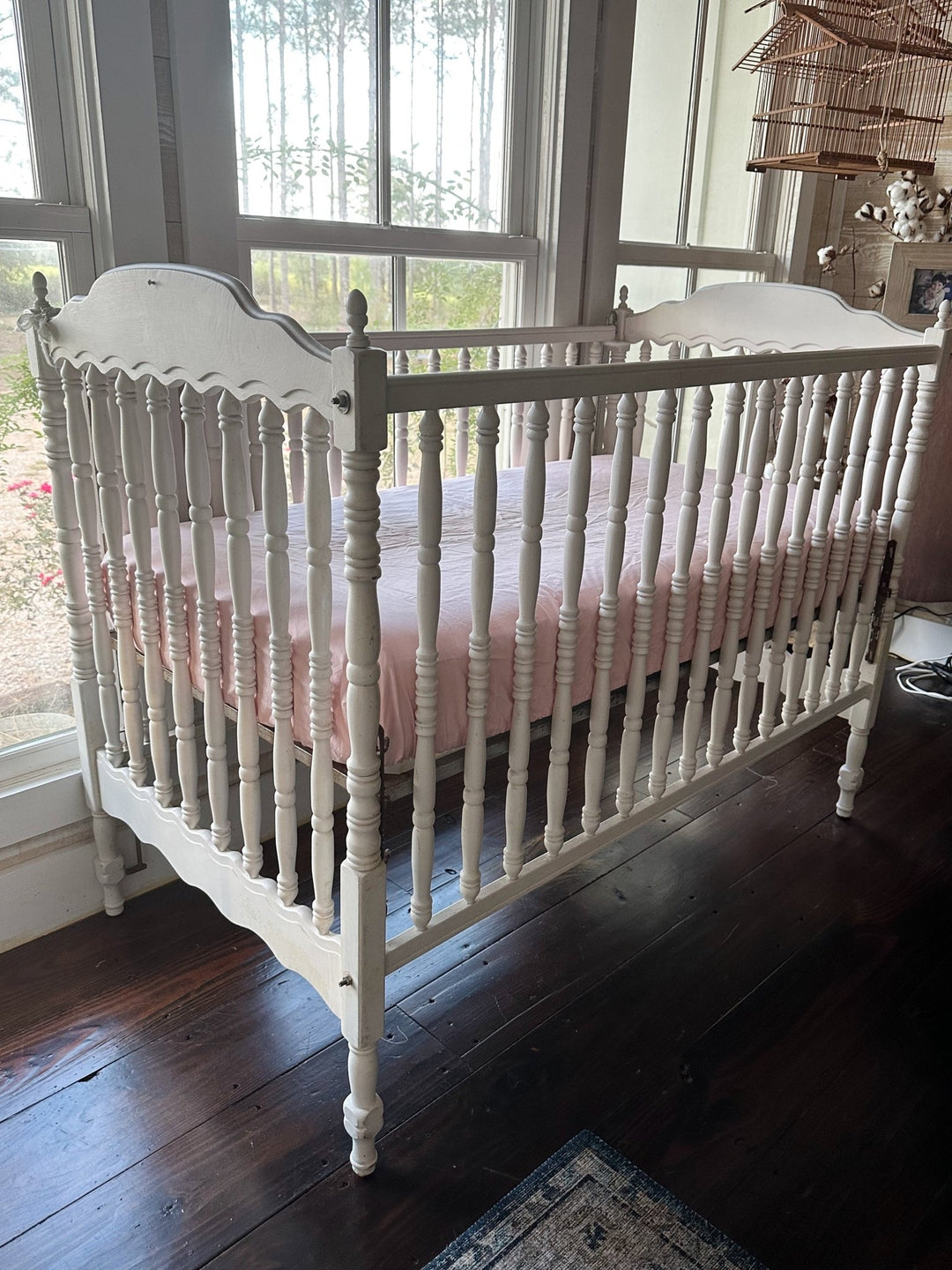 White spindle crib made up with a Sisters Pink fitted crib sheet beside tall windows, cotton stems and a hanging birdcage