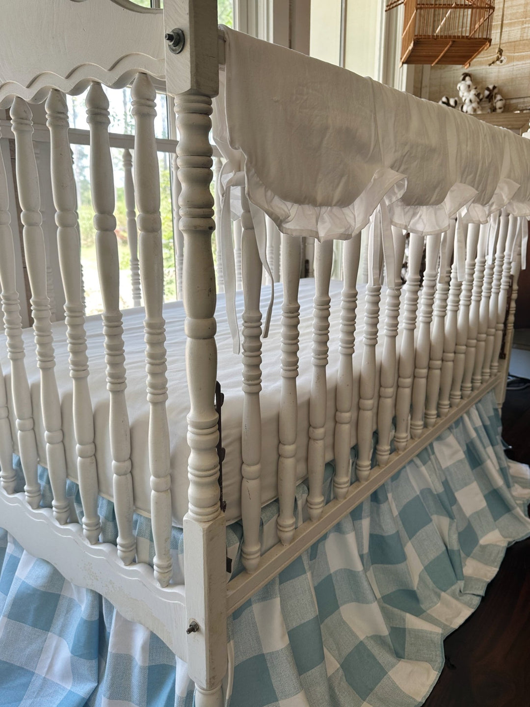 Side view of the white scalloped crib rail cover tied along the long top rail above a blue buffalo check skirt
