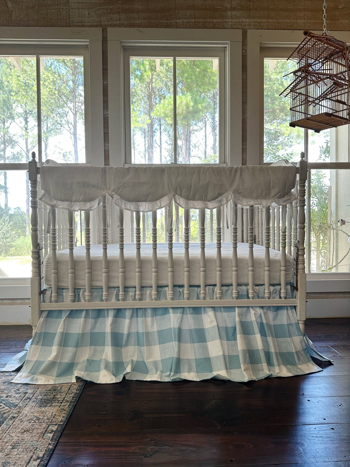 White ruffled scalloped crib rail cover and blue buffalo check farmhouse crib skirt on a white spindle crib