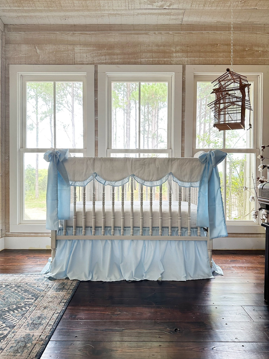 Heirloom blue and white crib bedding set styled beneath three windows in a pine-framed nursery