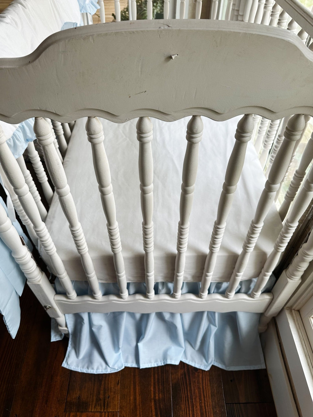 End view of the crib showing the white fitted sheet, heirloom blue skirt and the pillow resting at the side