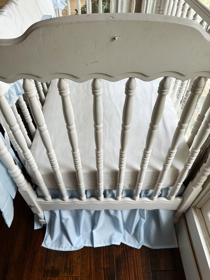 End view of the crib showing the white fitted sheet, heirloom blue skirt and the pillow resting at the side