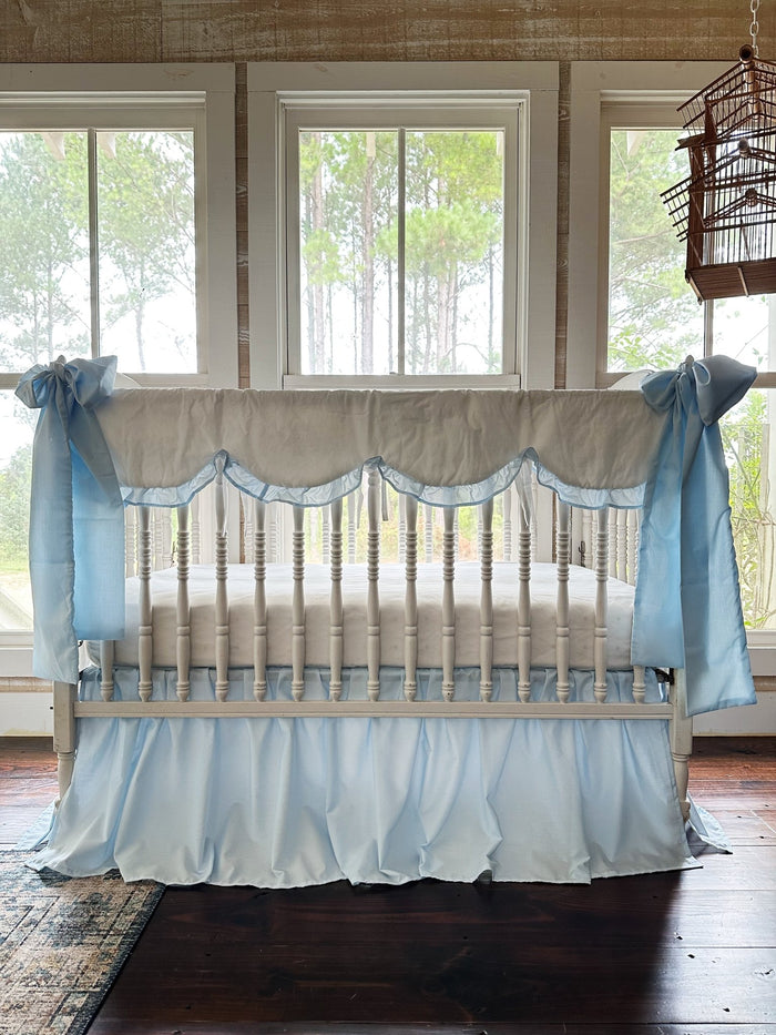 White scalloped crib rail cover with heirloom blue ruffle, blue farmhouse skirt and two large blue bows on a white spindle crib