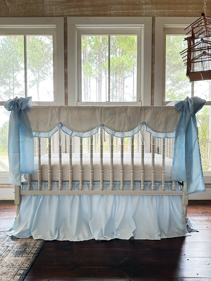White scalloped crib rail cover with heirloom blue ruffle, blue farmhouse skirt and two large blue bows on a white spindle crib
