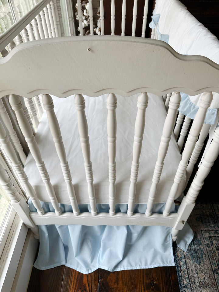 Looking down into the crib from the end over a white fitted sheet with blue skirt beneath