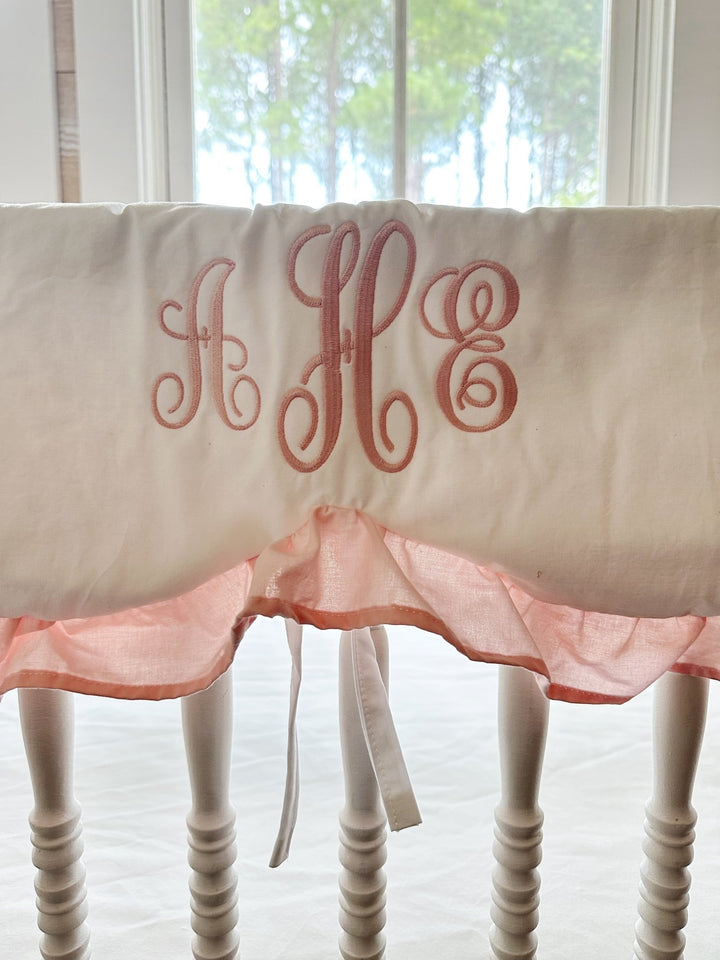 Close-up of a pink script monogram embroidered on a white scalloped crib rail cover