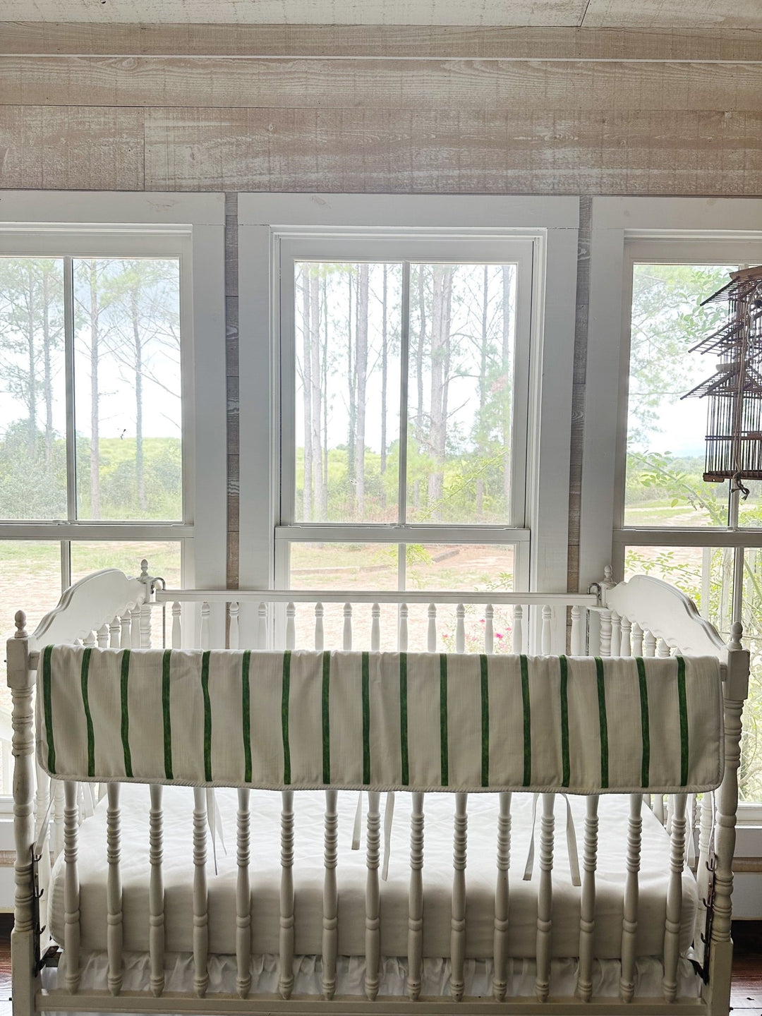 The striped rail cover on a crib in a pine-paneled nursery with windows and a hanging birdcage