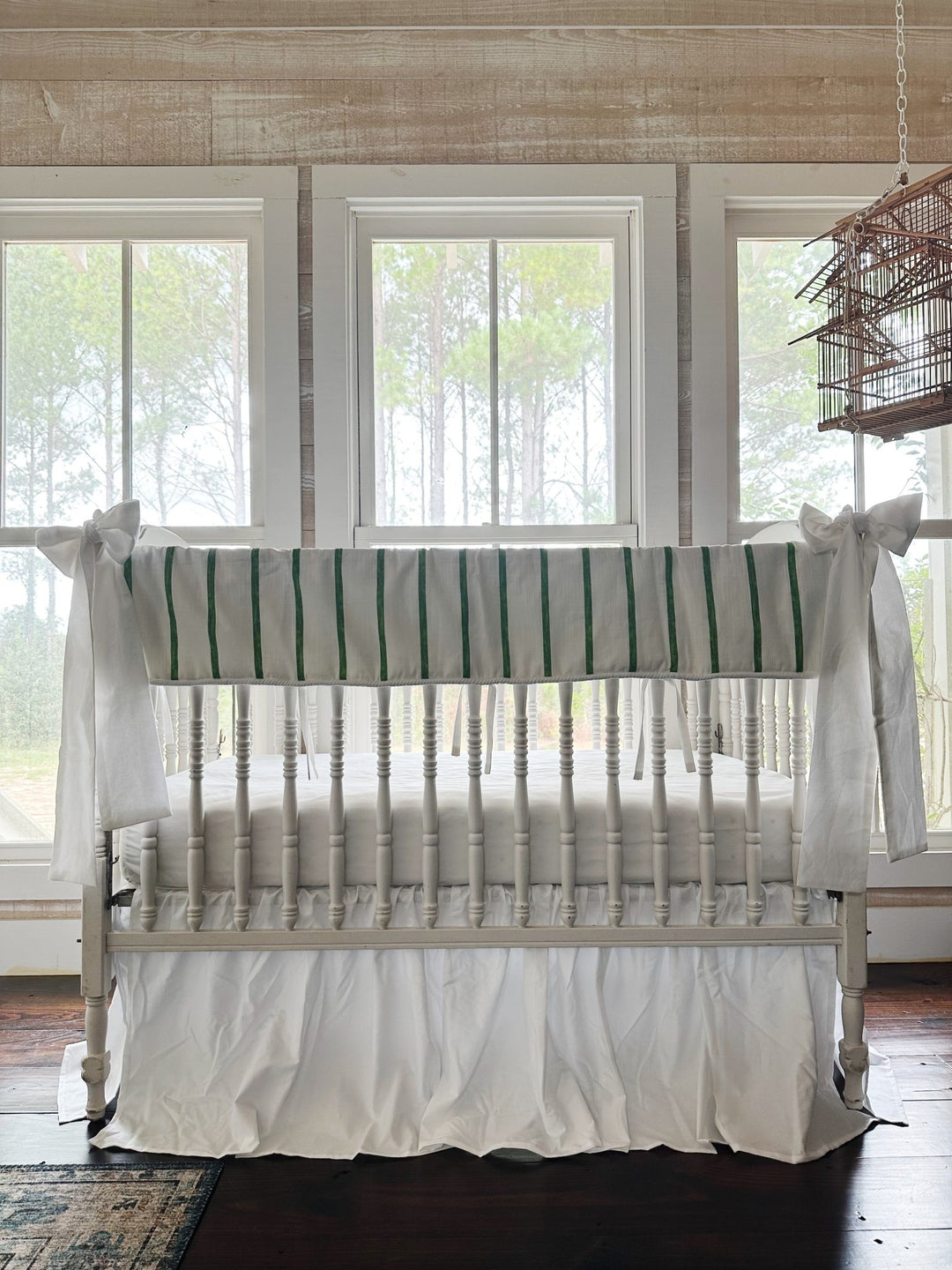Green and white striped tailored crib rail cover with a white farmhouse skirt and two white bows on a white spindle crib