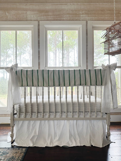 Green and white striped tailored crib rail cover with a white farmhouse skirt and two white bows on a white spindle crib