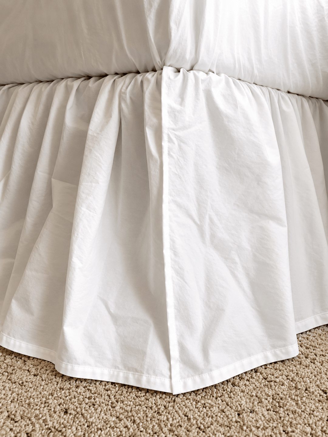 Close view of the white bed skirt corner over carpet, showing the gathered heading and the plain turned hem with no ruffle
